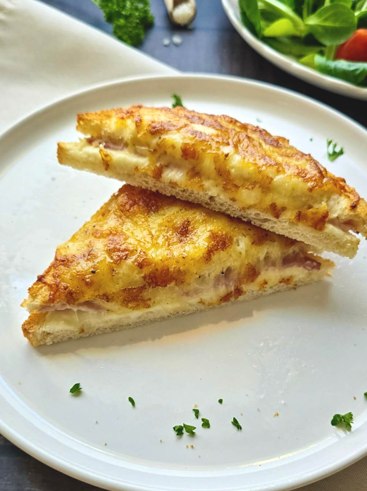 Un gros plan d'un croque-monsieur coupé en deux en diagonale, révélant un cœur fondant composé de jambon et de fromage coulant. Les triangles de pain sont superposés sur une assiette blanche. La croûte supérieure est intensément dorée et gratinée, parsemée de quelques éclats de persil frais. En arrière-plan, on distingue une salade verte croquante.