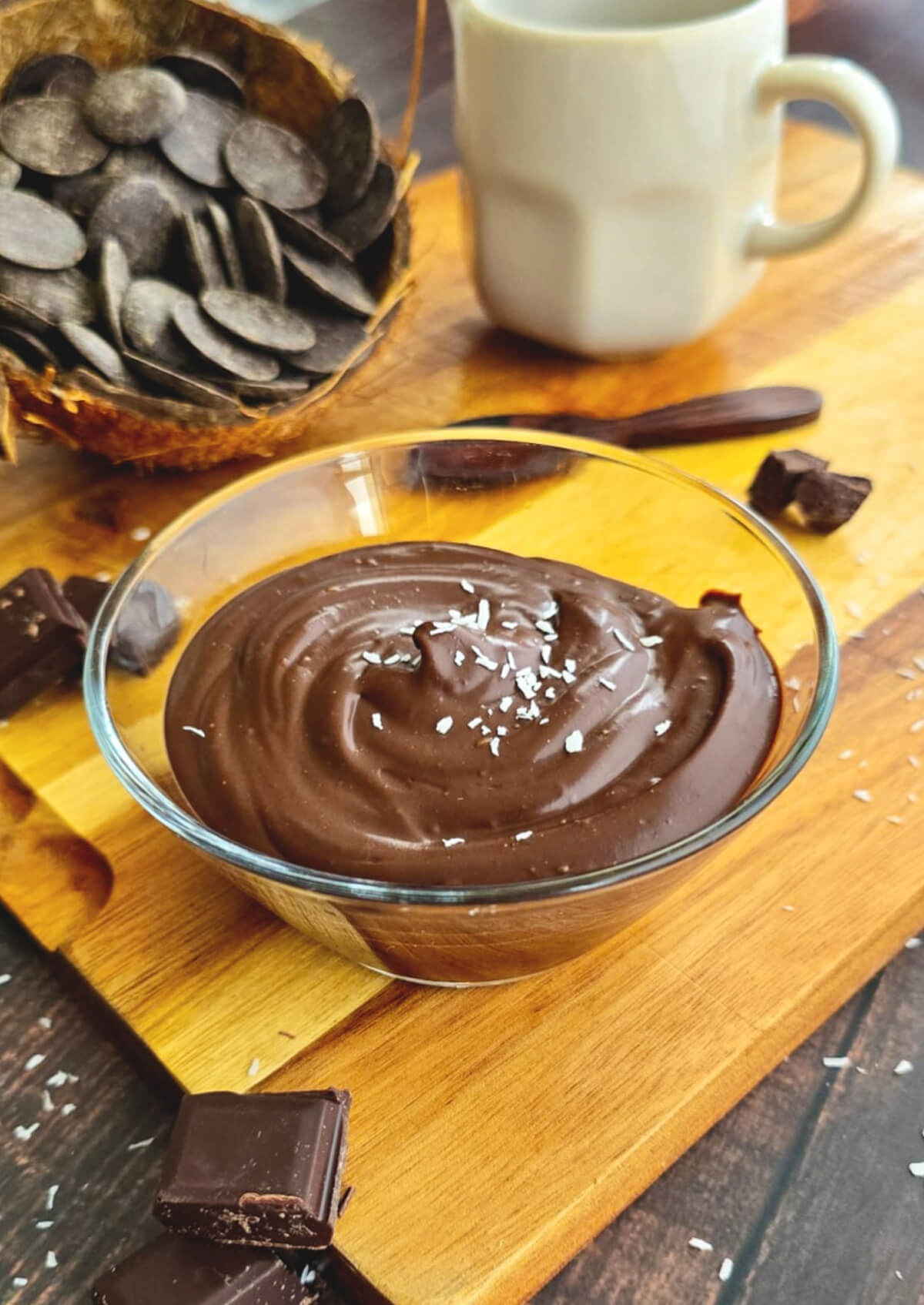 Crème dessert onctueuse au chocolat et lait de coco servie dans un bol en verre, saupoudrée de copeaux de noix de coco, présentée sur une planche en bois avec des carrés de chocolat noir et une coque de noix de coco remplie de pistoles de chocolat.