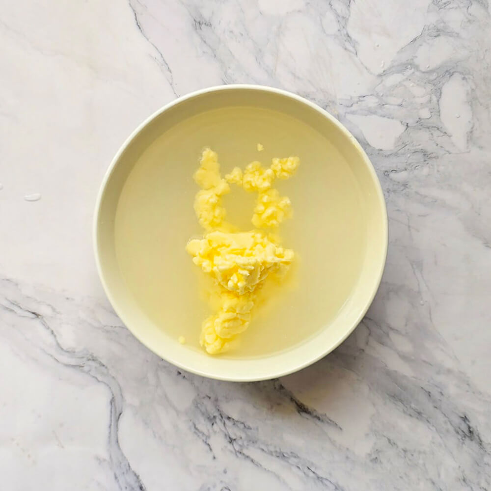 Une vue de dessus d'un bol blanc contenant des morceaux de beurre frais immergés dans de l'eau claire et très froide. L'eau est trouble autour des amas de beurre, montrant le processus d'élimination du petit-lait résiduel pour une meilleure conservation.