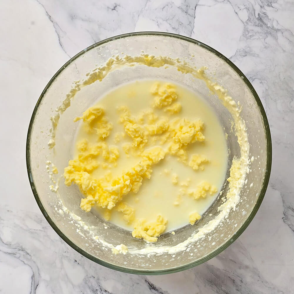 Une vue de dessus d'un bol en verre transparent où l'on voit clairement des grains de beurre jaunes flotter à la surface d'un liquide laiteux blanchâtre (le babeurre). Le liquide est trouble et les amas de beurre sont bien distincts.