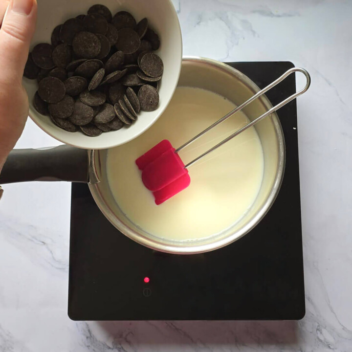 Action de verser des pistoles de chocolat noir pâtissier dans une casserole contenant un mélange de lait et de lait concentré chaud sur une plaque de cuisson.