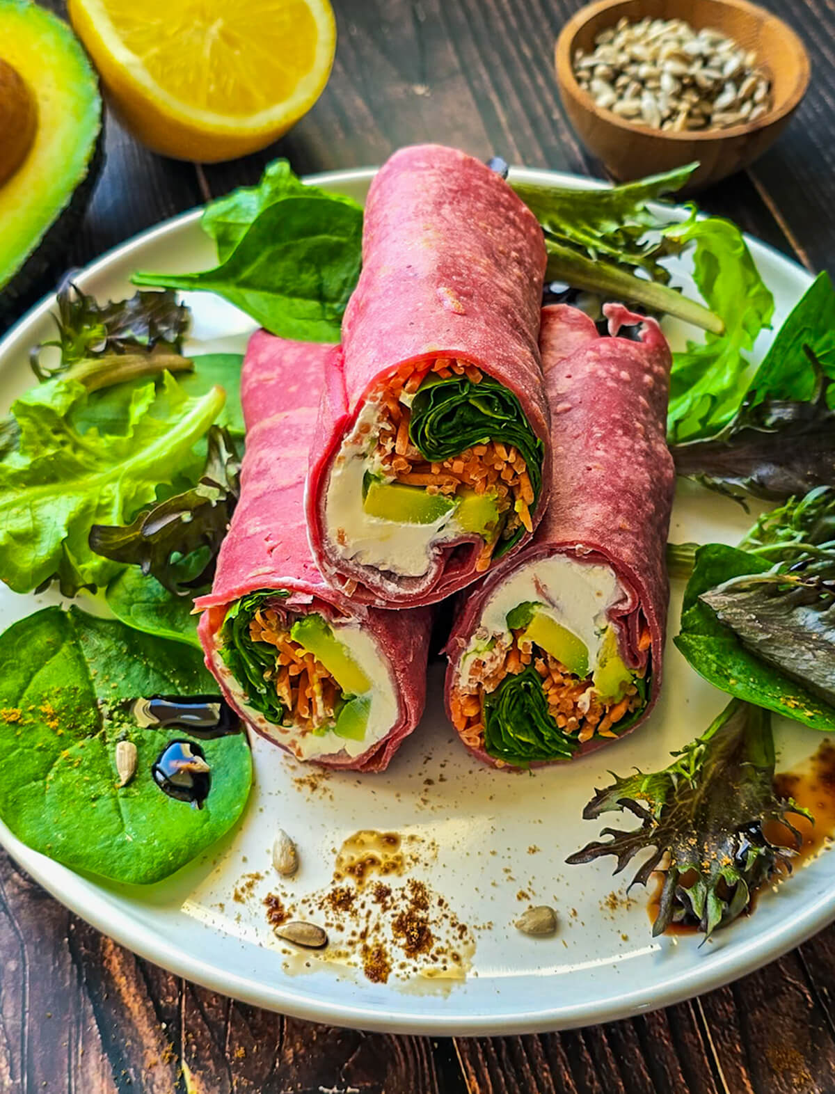 Trois wraps roses à la betterave coupés en deux et empilés sur une assiette, révélant une garniture fraîche de fromage de chèvre crémeux, d'avocat fondant, de carottes râpées et de jeunes pousses de chèvre. L'assiette est décorée de salade, de graines de tournesol et d'un filet de crème de balsamique sur un fond en bois sombre.