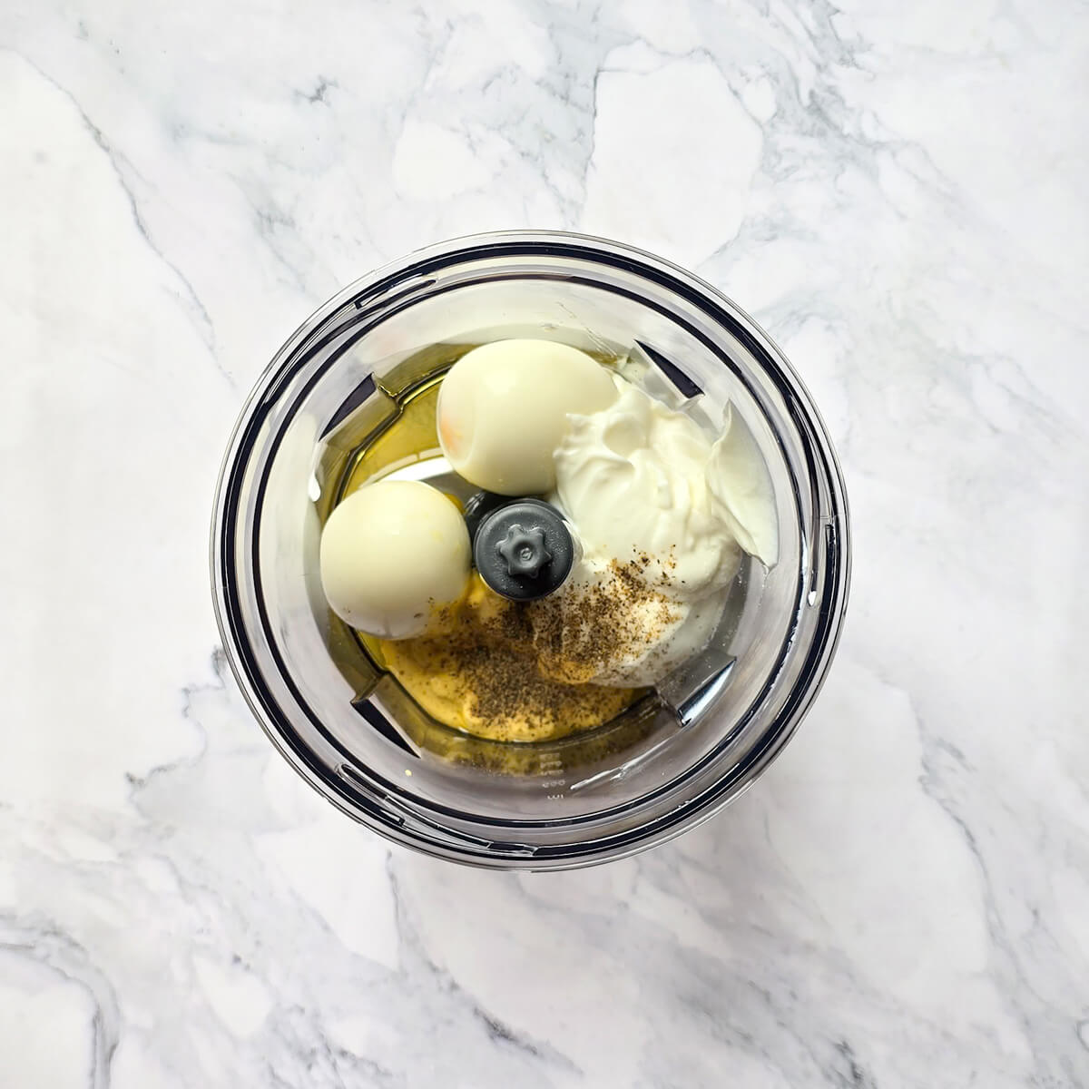 Ingrédients placés dans la cuve d'un mixeur : œufs durs écalés, Skyr, moutarde, huile d'olive et poivre, prêts à être mixés pour une sauce onctueuse.