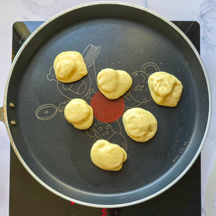 Vue de dessus de six petits tas de pâte à pancake healthy à la banane en train de cuire sur une poêle antiadhésive T-fal noire, posée sur une plaque de cuisson à induction. Les bords commencent à figer.
