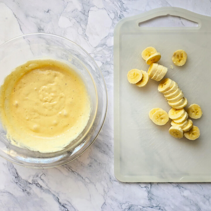 Plan de travail en marbre avec un bol en verre contenant la pâte à pancake liquide et jaune, à côté d'une planche à découper blanche où repose une banane mûre coupée en rondelles régulières.