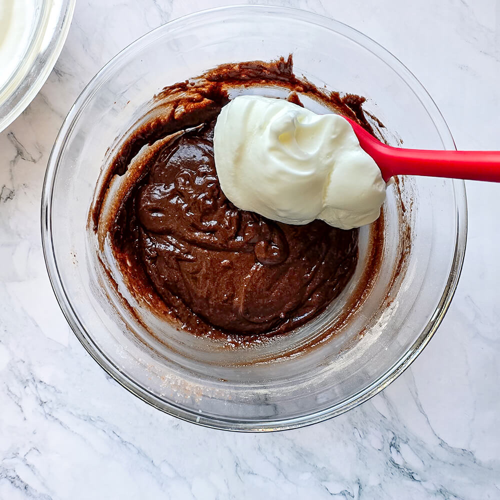 Gros plan d'une spatule rouge incorporant délicatement une meringue blanche et mousseuse dans une pâte à gâteau au cacao onctueuse, dans un grand saladier en verre. La texture de la pâte est visible et prête à être transférée dans le moule pour la cuisson.