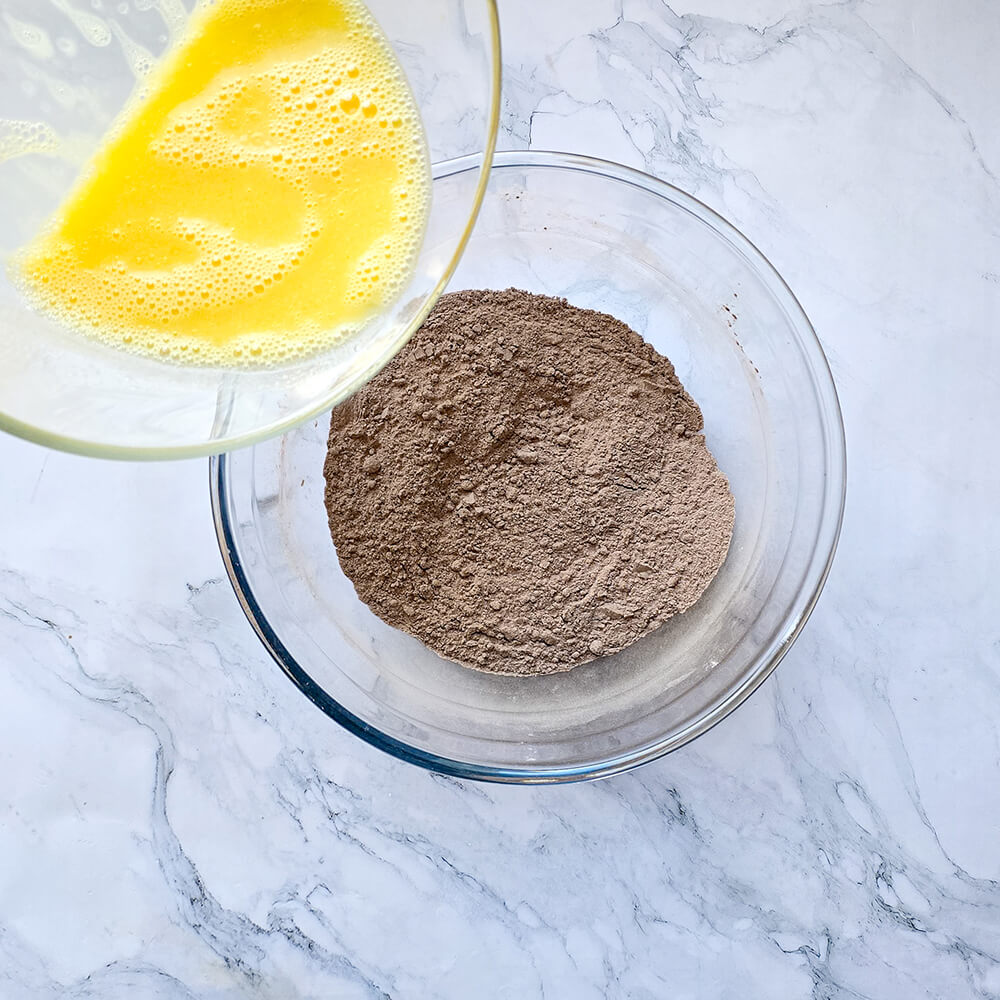 Mains versant un bol d'œufs battus jaunes et mousseux dans un grand saladier en verre contenant le mélange sec (farine et cacao) d'un chiffon cake au cacao.