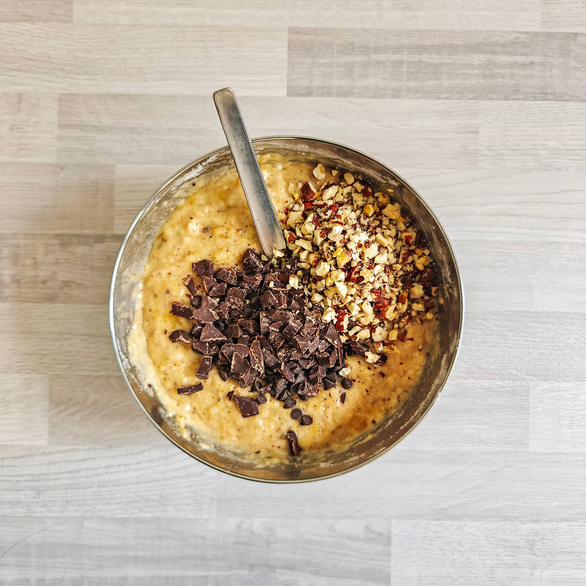 Vue de dessus d'une pâte à cake épaisse dans un bol en acier, sur laquelle sont déposés un généreux tas de pépites de chocolat noir concassées et un tas de noisettes entières hachées, avec une cuillère insérée dans le mélange.