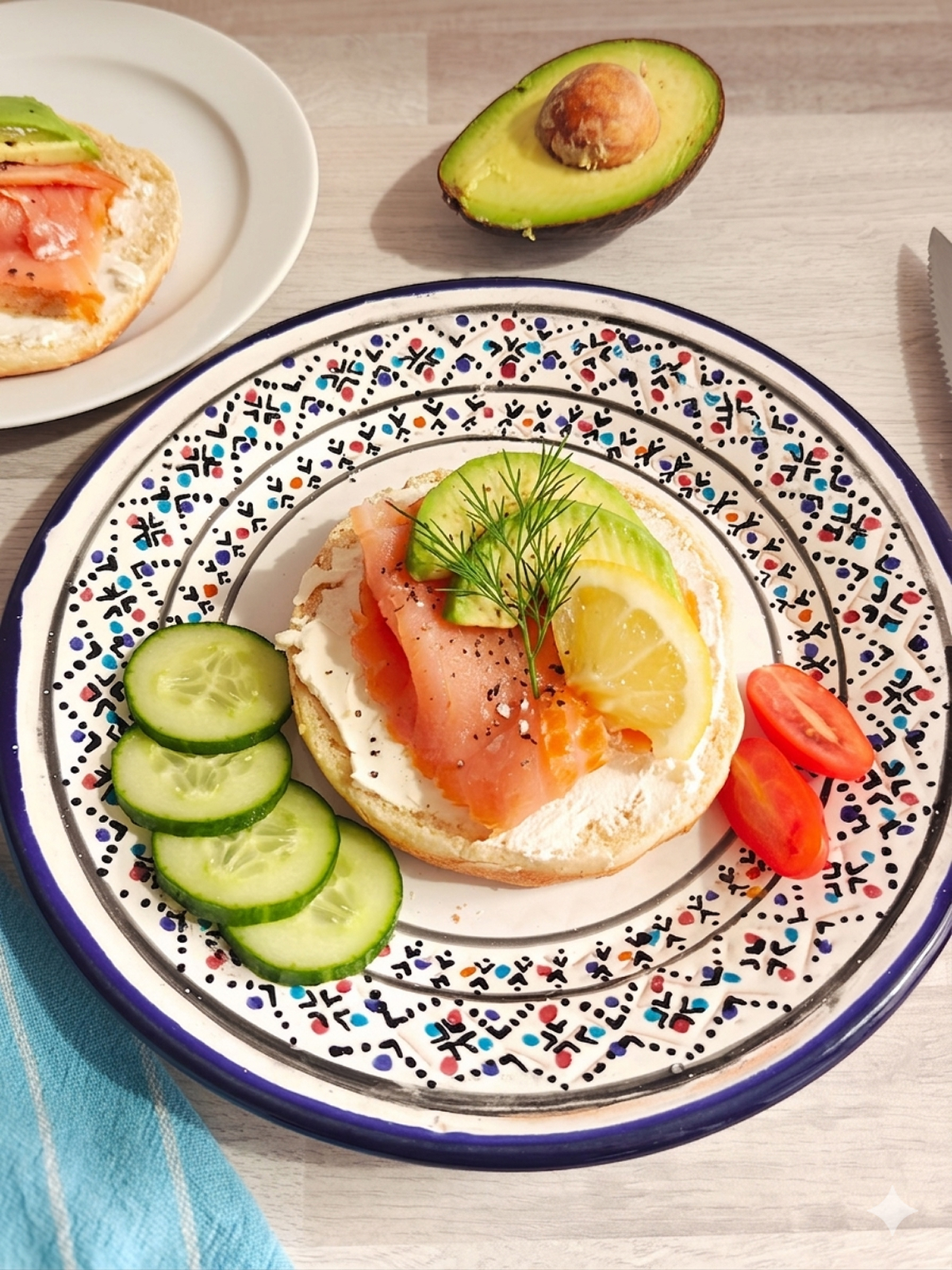 Un demi-bagel garni de fromage frais, d'une tranche généreuse de saumon fumé et de lamelles d'avocat. L'assiette est décorée d'une branche d'aneth, d'une tranche de citron, de rondelles de concombre et de tomates cerises.