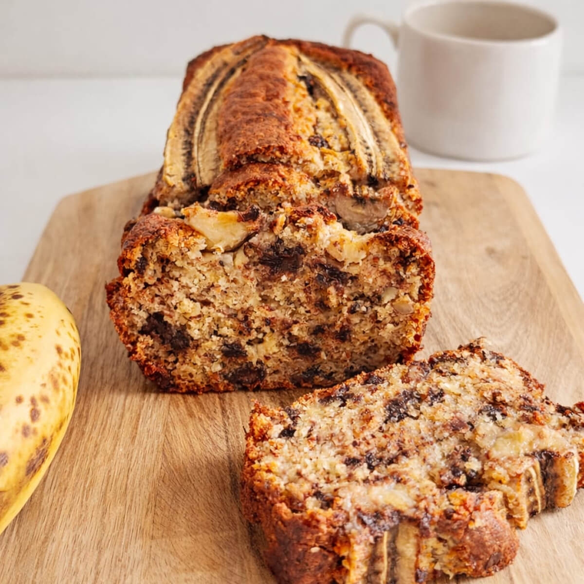 Un cake à la banane (banana bread) tranché sur une planche en bois, révélant une mie moelleuse aux pépites de chocolat fondu et éclats de noisettes, décoré d'une banane entière sur le dessus.