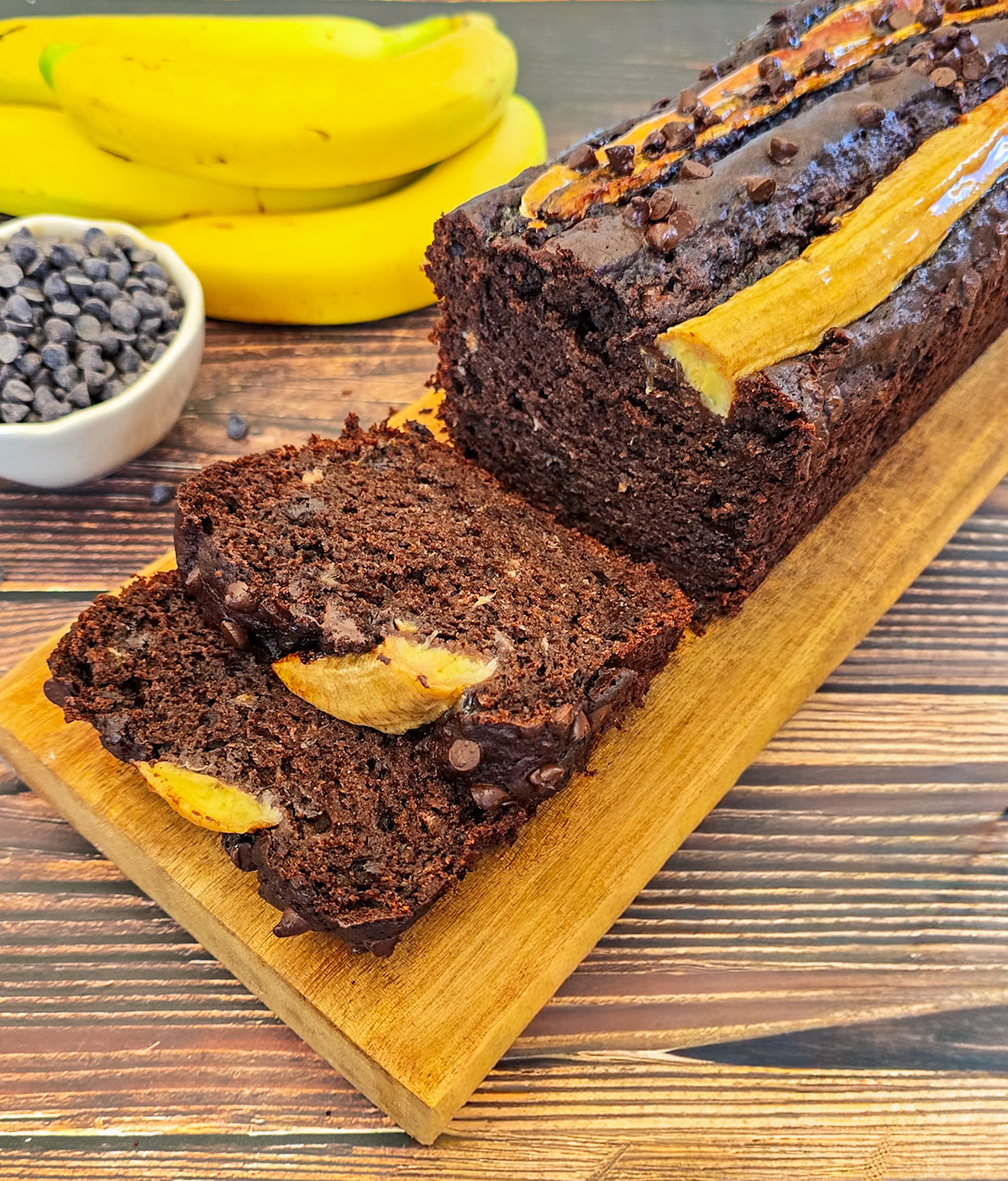 Gros plan sur des tranches de banana bread au chocolat sur une planche de bois, bananes et pépites de chocolat en arrière.