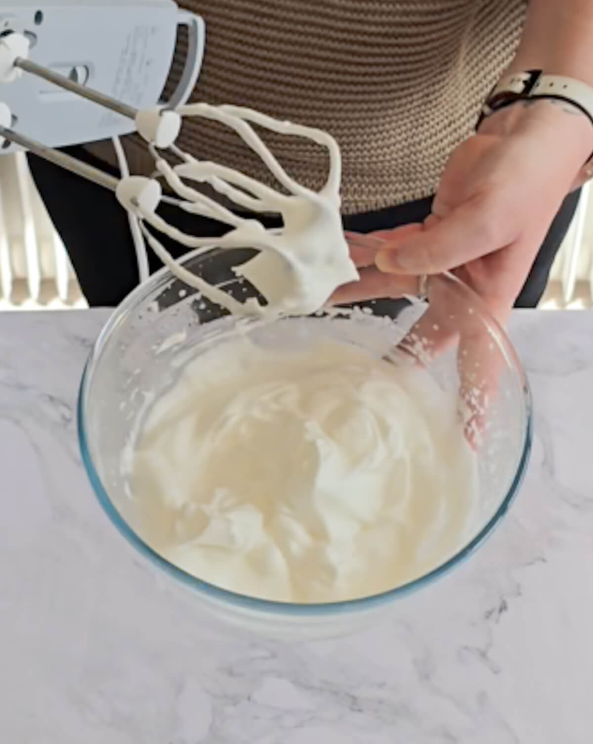 Une main tenant un saladier de blancs en neige pas assez montés et une autre main tenant un fouet électrique.