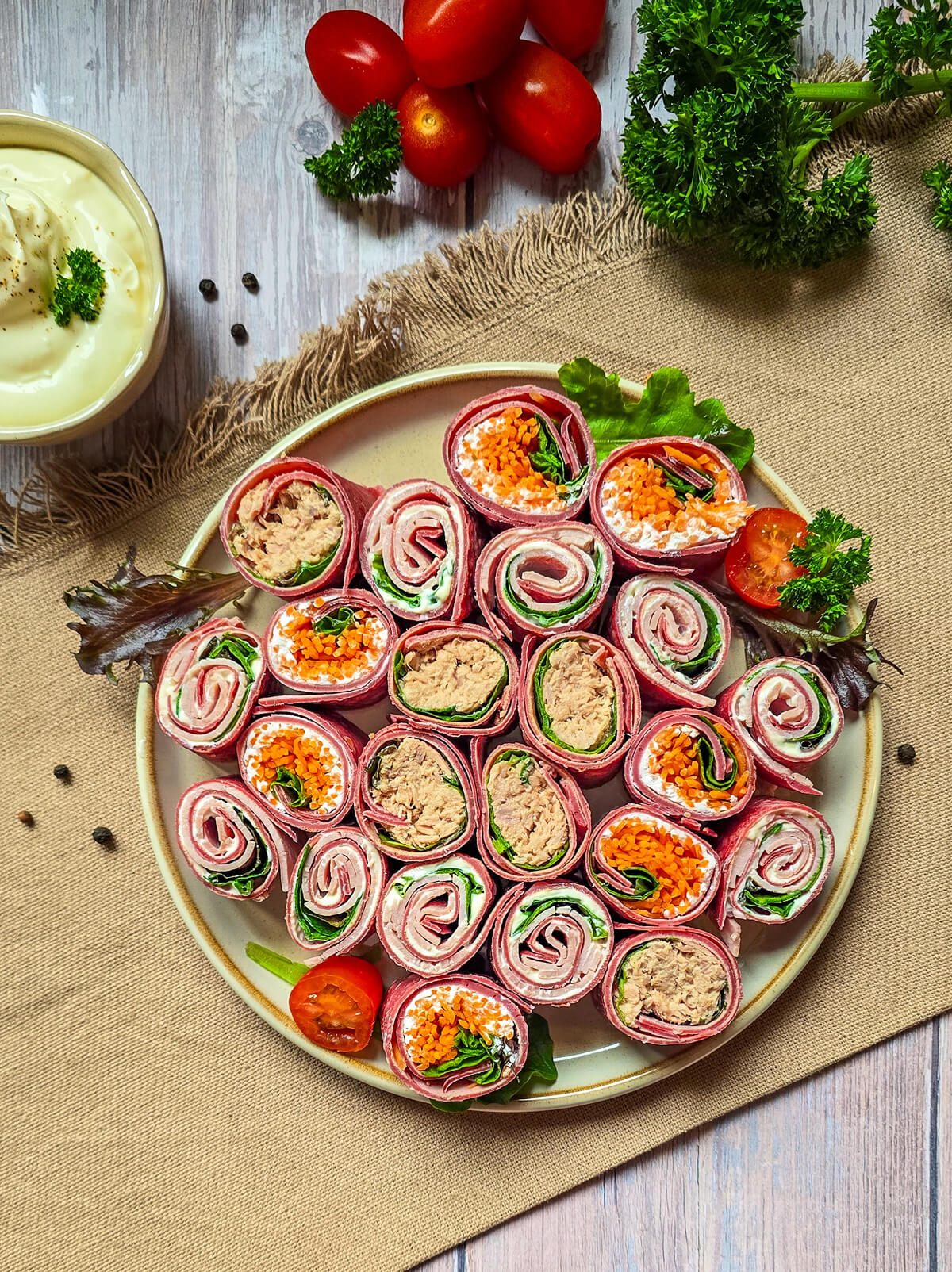 Gros plan sur une assiette blanche rectangulaire présentant trois bouchées de wraps à la betterave (makis). On distingue clairement une garniture au thon, une autre aux carottes râpées et une troisième au fromage frais. L'assiette est décorée d'une demi-tomate cerise et d'une feuille de chêne, avec une fourchette en argent gravée "Cuisine & Création". Le plateau principal est flou en arrière-plan.