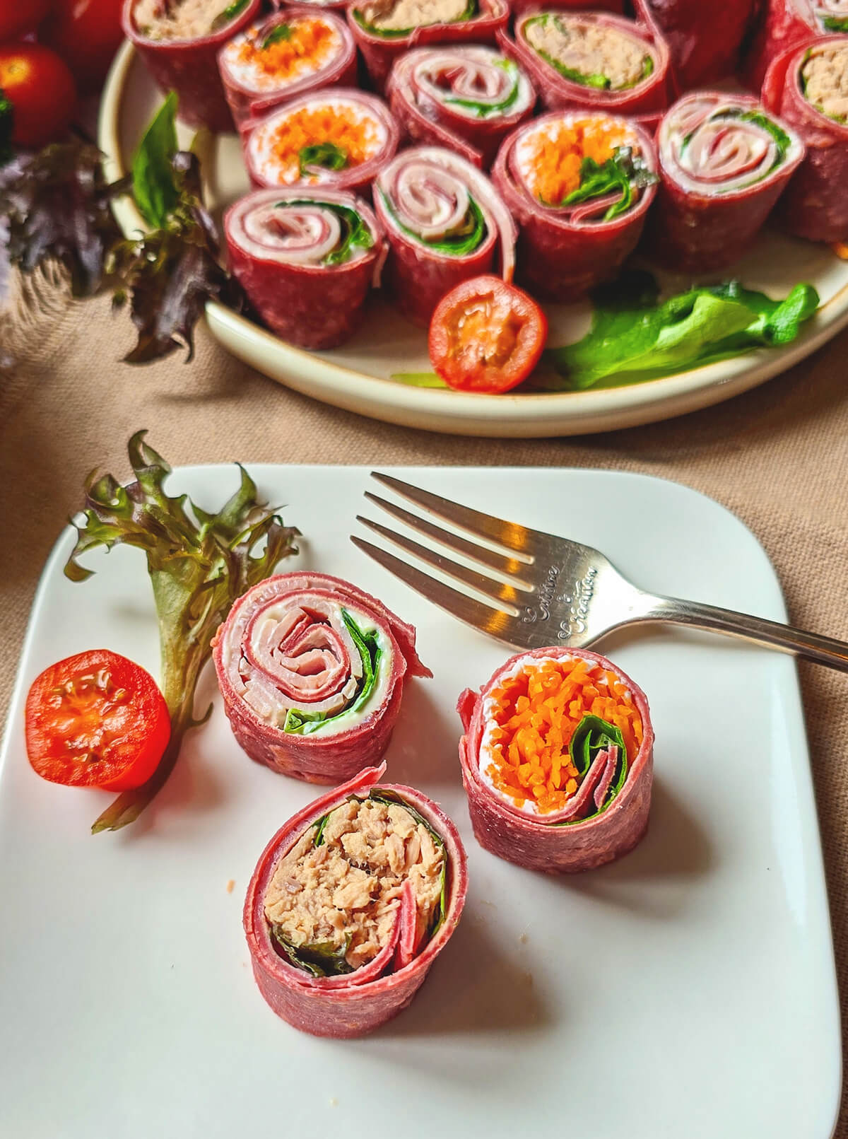 Vue de dessus d'un grand plateau rempli de bouchées de wraps à la betterave (makis healthy). Elles sont garnies de thon, carottes râpées, salade et fromage frais. Sur le côté, on aperçoit un bol de mayonnaise skyr, des tomates cerises et du persil, le tout posé sur une nappe en jute et une table en bois clair.