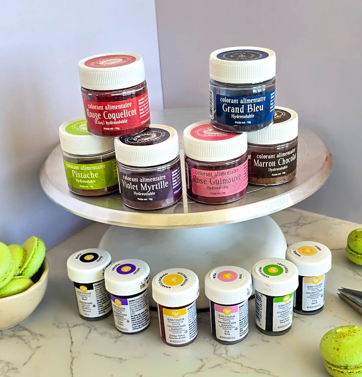 Une sélection de colorants alimentaires pour macarons : des pots de colorants hydrosolubles en poudre sur un support rotatif (rouge, bleu, violet, vert) et des petits flacons de colorant en gel Wilton au premier plan, à côté de macarons verts.