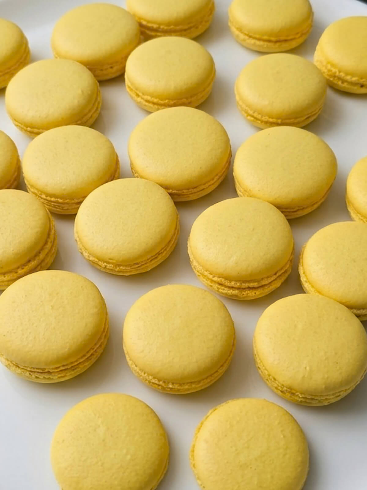 Plusieurs rangées de coques de macarons d'un jaune vif et uni, lisses et brillantes, refroidissant sur un plan de travail blanc.