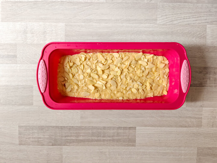 Vue de dessus d'un moule à cake rectangulaire en silicone rose vif, posé sur un plan de travail en bois clair. À l'intérieur, la pâte à banana bread est étalée uniformément et généreusement recouverte d'amandes effilées prêtes pour la cuisson.