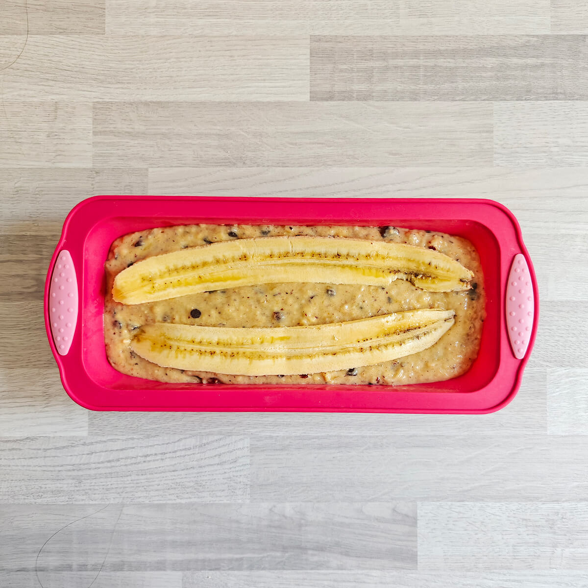 Vue de dessus d'un moule à cake en silicone rose vif rempli de pâte, décoré de deux demi-bananes entières coupées dans la longueur et caramélisées, prêt à être enfourné.