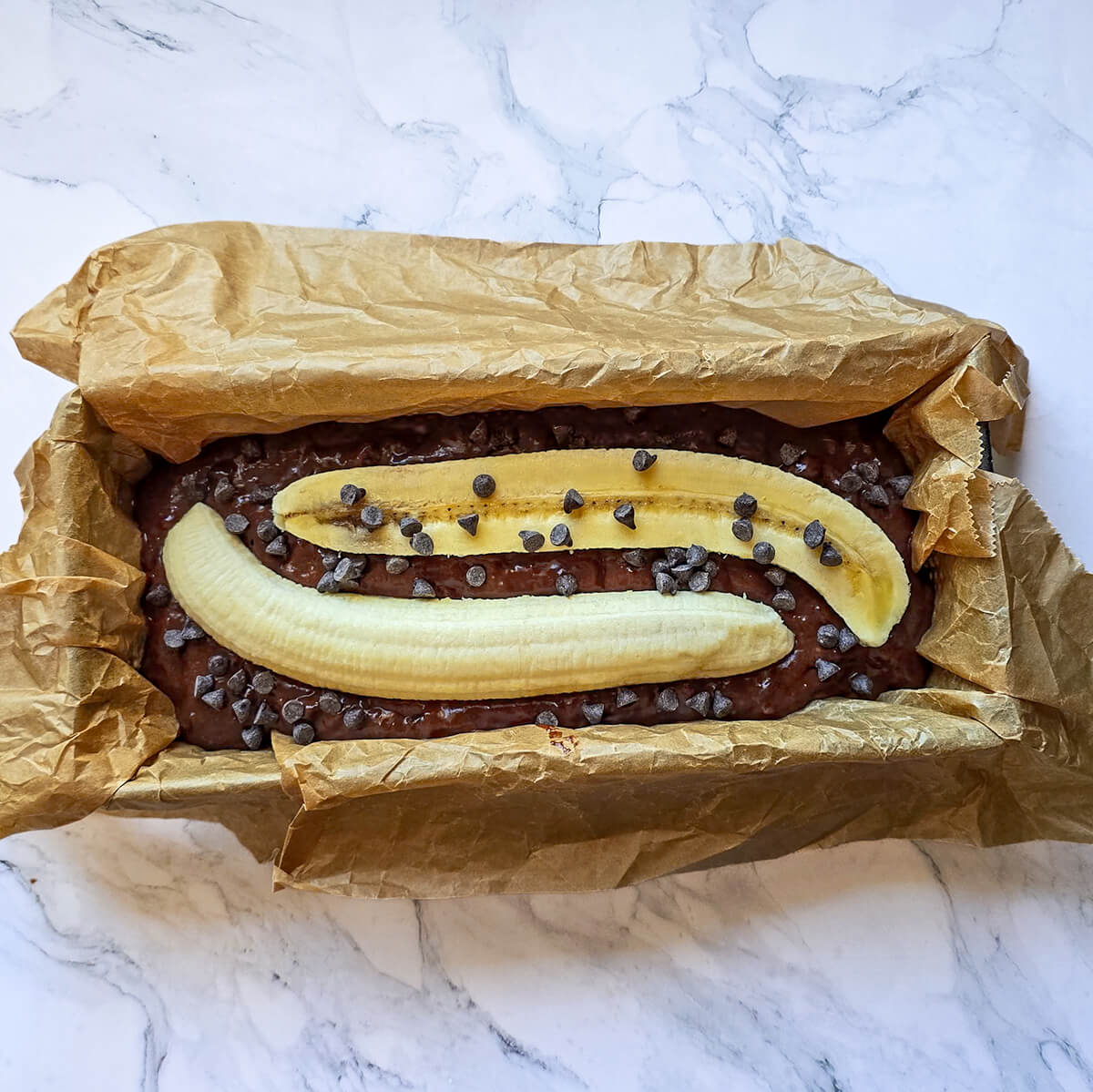 Un moule à cake tapissé de papier sulfurisé est rempli de pâte au chocolat. Deux moitiés de banane et des pépites de chocolat décorent le dessus avant la cuisson.