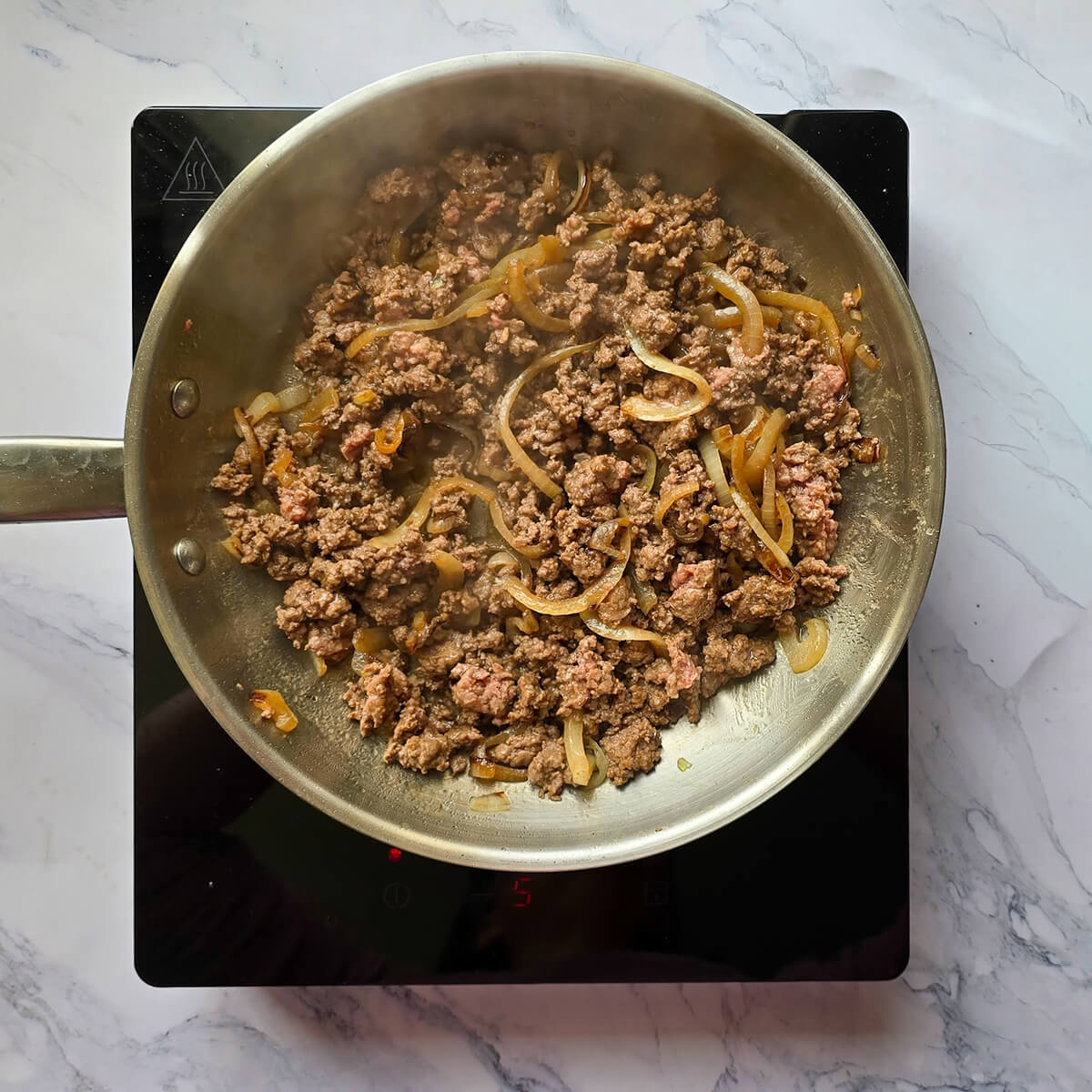 Viande de bœuf hachée et oignons émincés en train de revenir dans une poêle en inox sur une plaque à induction.