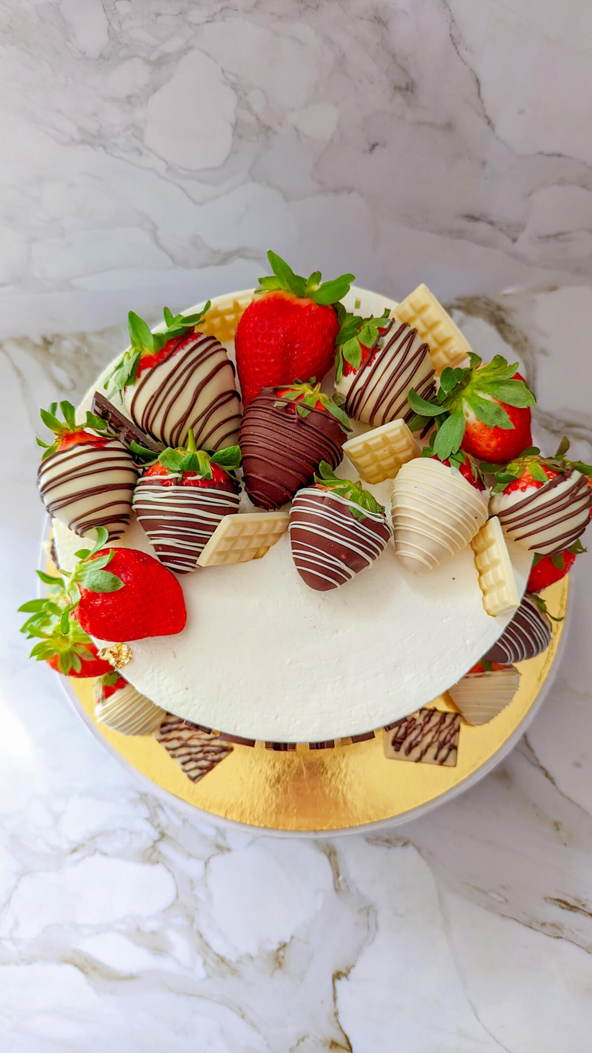 Vue de dessus d'un layer cake recouvert d'une chantilly mascarpone lisse, généreusement décoré de fraises fraîches entières, de fraises enrobées de chocolat blanc et de mini tablettes de chocolat blanc.