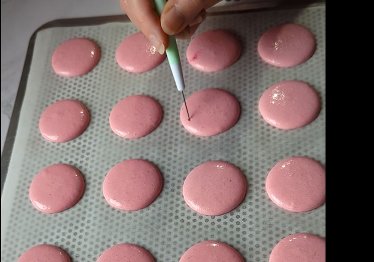 Une main utilisant une pointe fine pour percer délicatement les bulles d'air à la surface de coques de macarons roses pochées sur un tapis de cuisson perforé.
