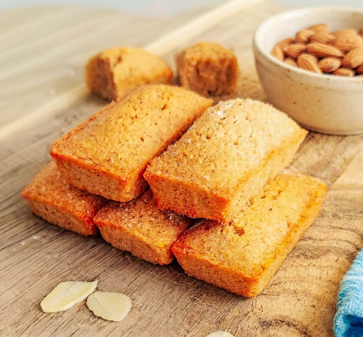 Gros plan sur une pile de financiers aux amandes dorés posés sur une planche en bois, avec un bol d'amandes entières en arrière-plan et des amandes effilées au premier plan.