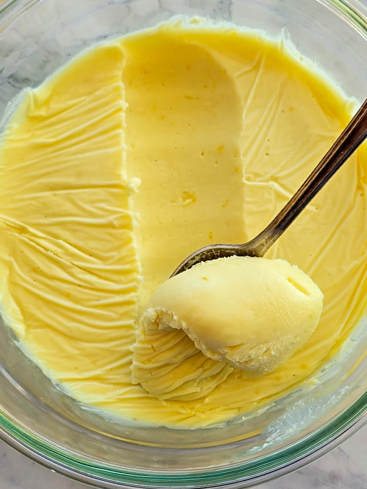 Une cuillère en argent prélevant une noisette généreuse de ganache au citron, jaune vif dans un bol en verre. La surface montre l'empreinte du film fraîcheur après refroidissement.