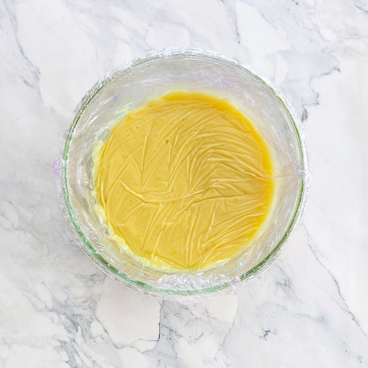 Ganache au citron jaune vif dans un bol en verre, recouverte de film fraîcheur au contact pour éviter qu'une croûte ne se forme, prête pour la réfrigération.
