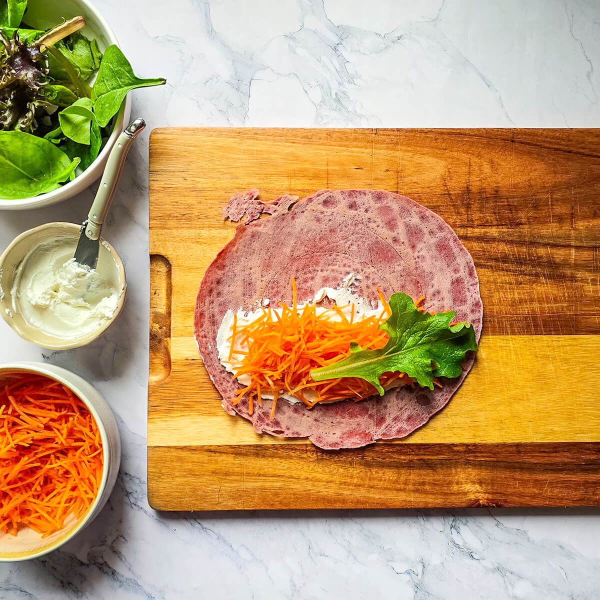 Vue de dessus d'une galette de wrap à la betterave maison posée sur une planche en bois. Elle est garnie au centre d'une bande de fromage frais tartiné, surmontée de carottes râpées orange vif et d'une feuille de laitue verte. Des bols d'ingrédients entourent la planche sur un plan de travail en marbre.