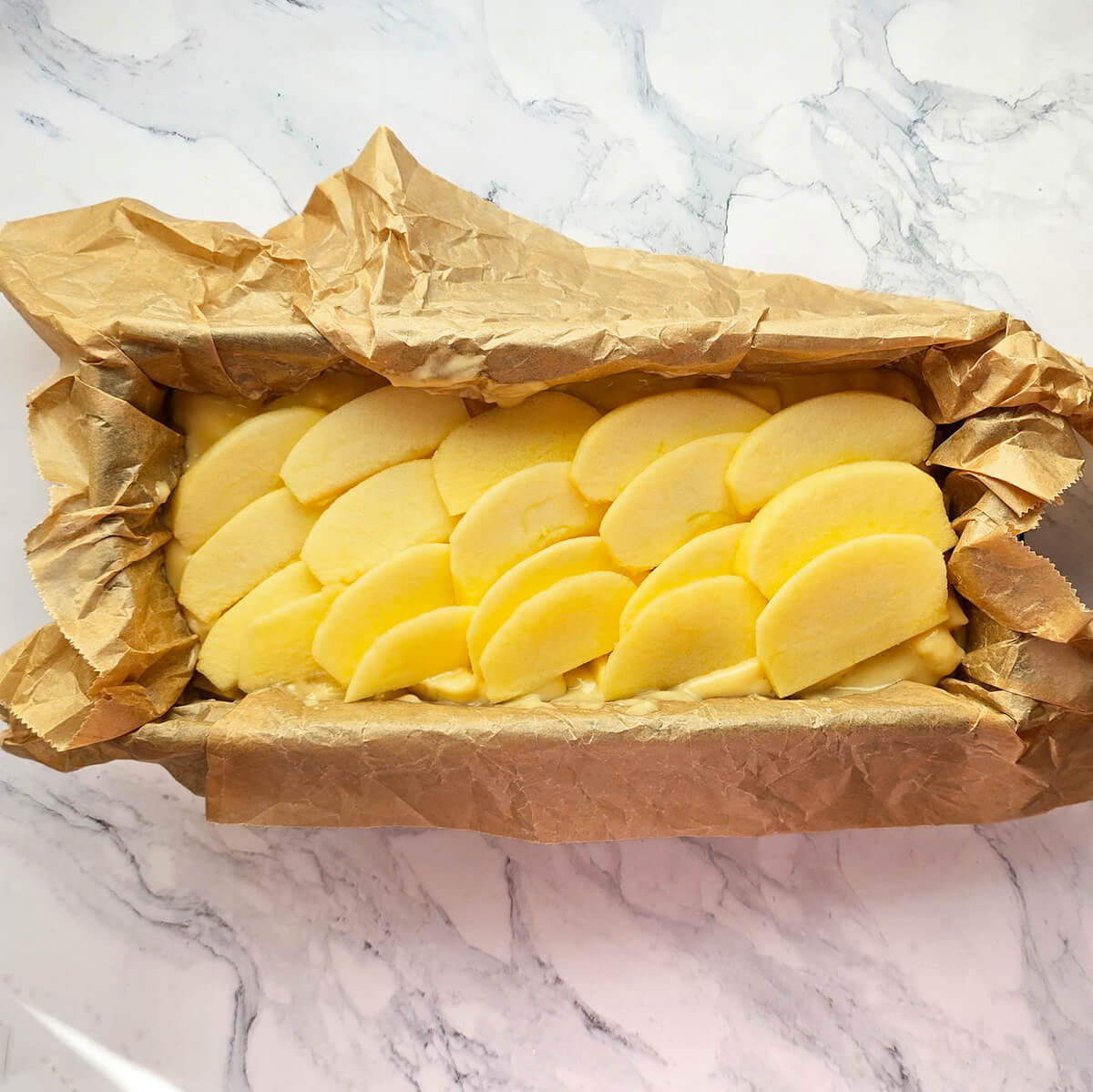 Vue de dessus d'un moule à cake tapissé de papier sulfurisé froissé, rempli de pâte et recouvert de lamelles de pommes rangées en rosace, prêt à être enfourné sur un plan de travail en marbre.