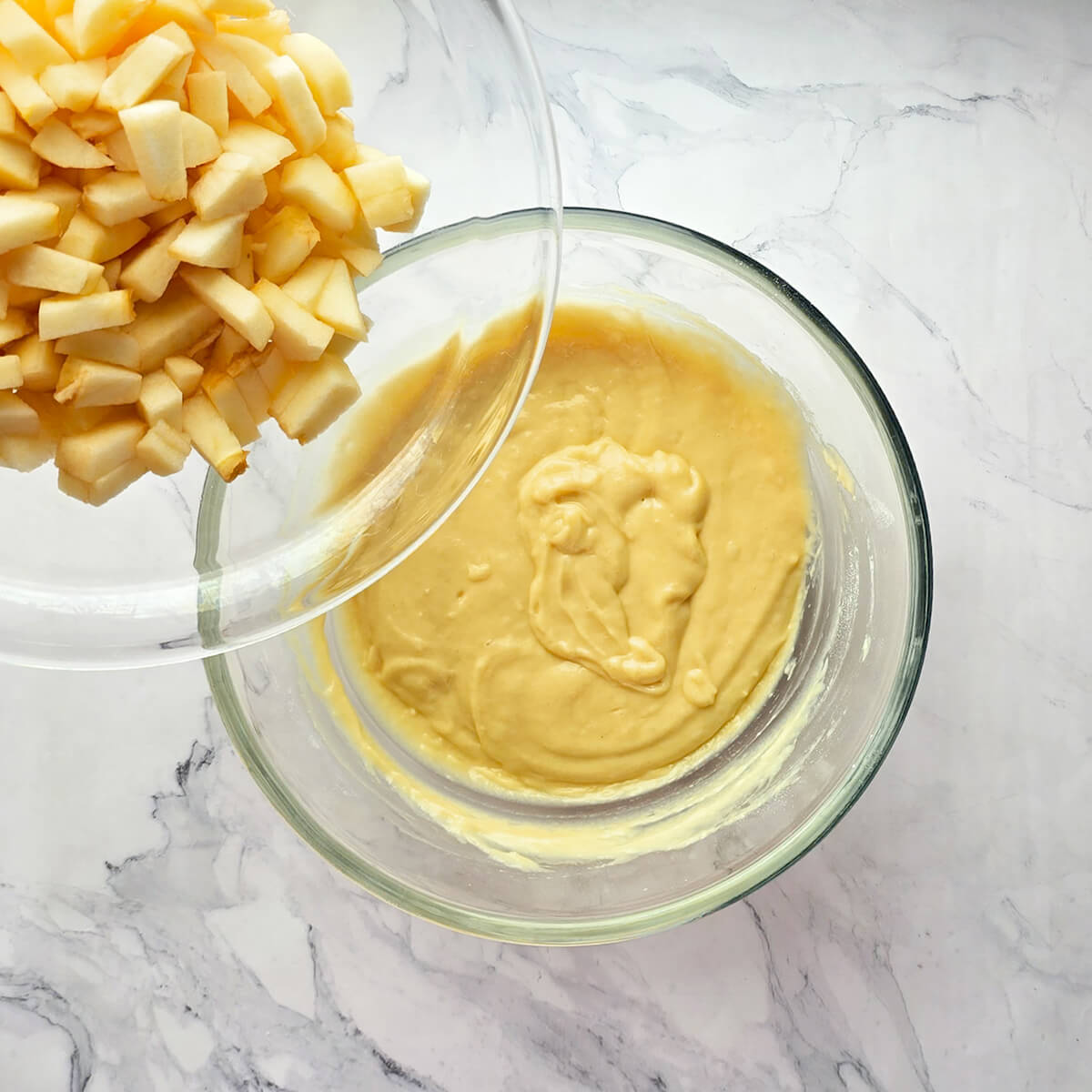 Vue de dessus d'un bol en verre contenant une pâte à gâteau lisse et jaune clair, dans lequel une main verse des dés de pommes fraîches depuis un autre bol.
