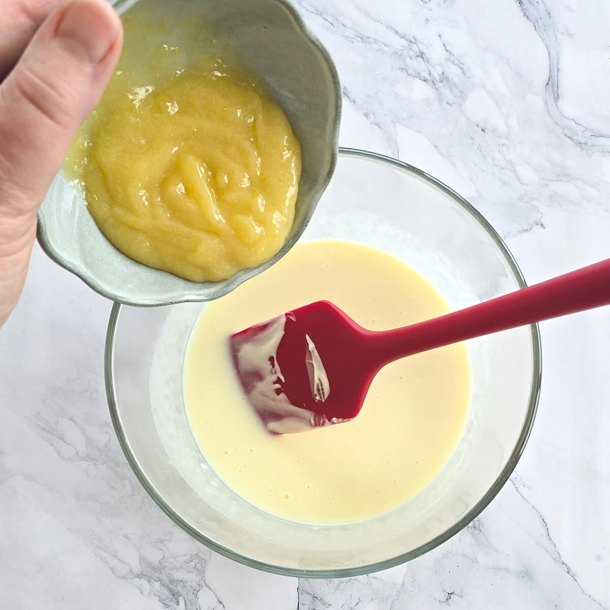 Une main verse du lemon curd onctueux d'un petit bol vert dans une base de chocolat blanc fondu dans un grand bol en verre pour faire une ganache.