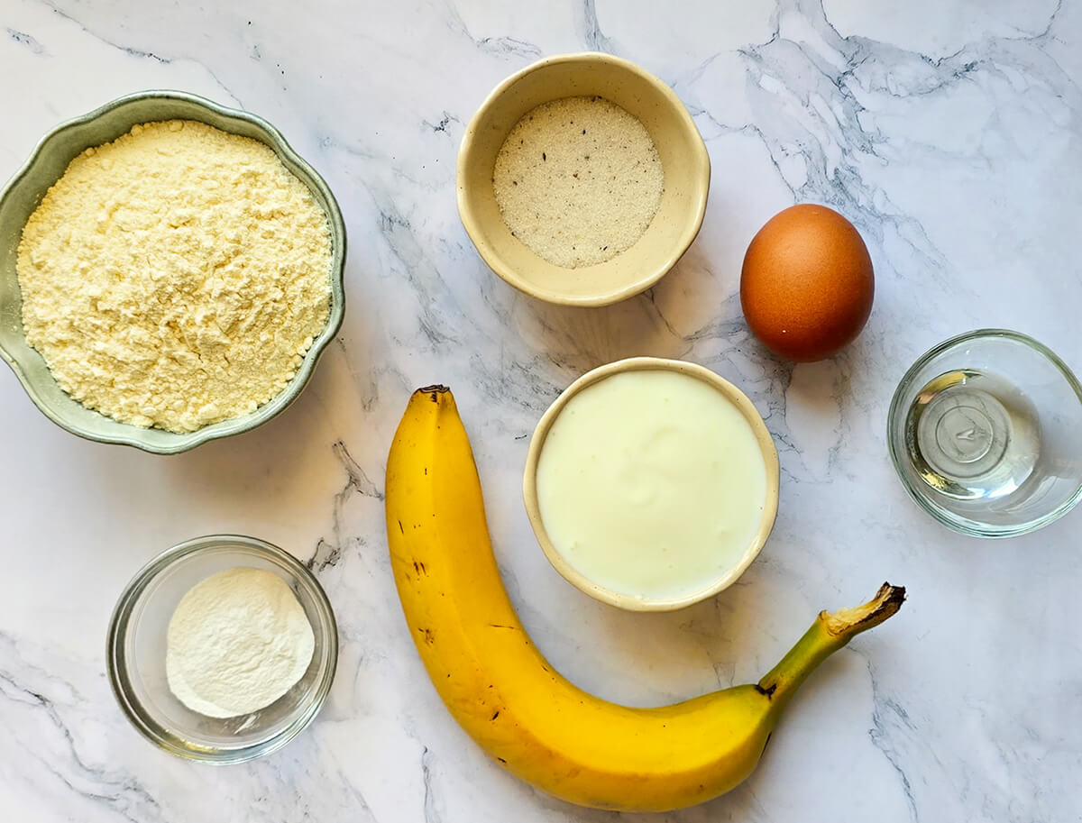 Vue de dessus des ingrédients pour préparer des pancakes à la banane healthy : farine de sarrasin, œuf, banane mûre, yaourt nature, sucre, poudre à lever et huile de coco, disposés dans des petits bols sur un plan de travail en marbre blanc.
