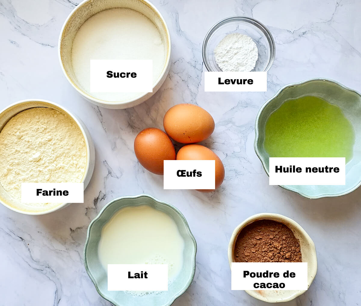 Des ingrédients pour préparer une recette de chiffon cake au cacao moelleuse, sur une surface blanche marbrée.