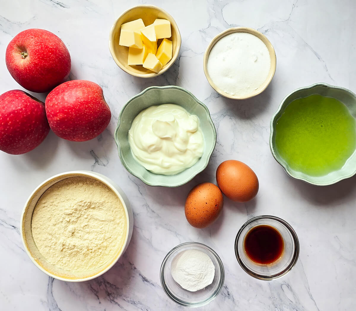 Vue de dessus des ingrédients pour un gâteau au yaourt aux pommes disposés sur un plan de travail en marbre blanc : trois pommes rouges, un bol de farine, un bol de yaourt, deux œufs, du sucre, de l'huile, des dés de beurre, de la levure et de l'extrait de vanille.