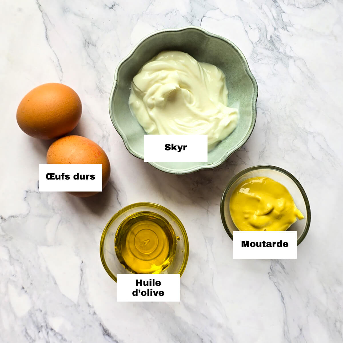 Vue de dessus des ingrédients pour une mayonnaise légère : deux œufs durs, un bol de Skyr, un ramequin d'huile d'olive et de la moutarde sur un plan de travail en marbre blanc.