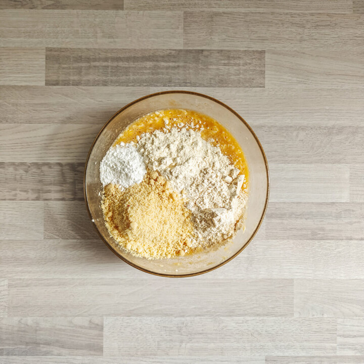 Une vue plongeante d'un bol en verre contenant un mélange humide (bananes écrasées et œufs). Sur le dessus, trois tas distincts d'ingrédients secs ont été ajoutés : de la farine bise, de la poudre d'amandes (plus jaune) et une petite dose de levure chimique blanche.