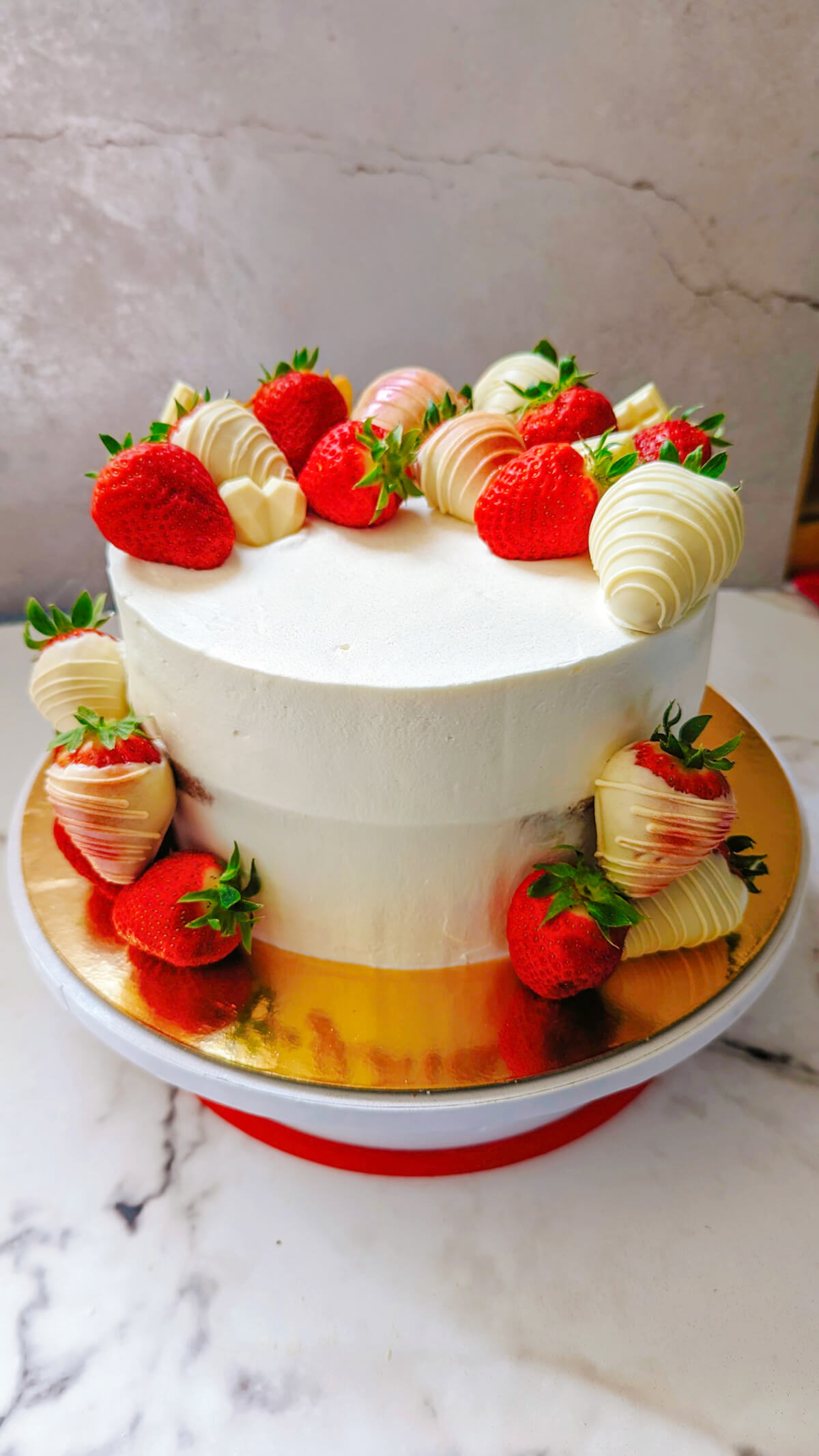 Un layer cake fraise mascarpone entier présenté sur un plat doré et marbré, avec un lissage parfait à la crème mascarpone et une décoration de fraises fraîches et chocolat blanc tout autour de la base et du dessus.
