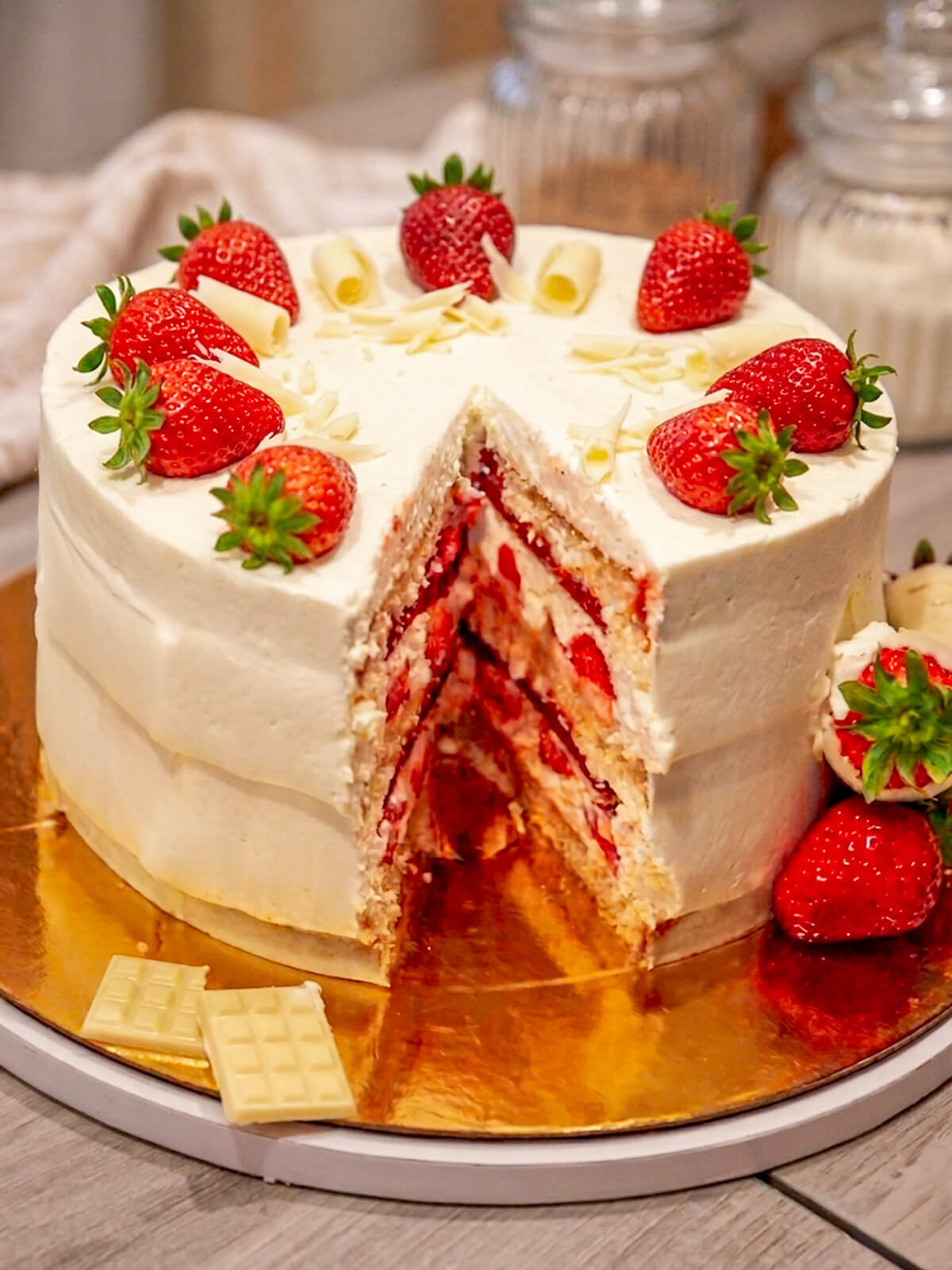 Une part découpée d'un layer cake fraise vanille sur un support doré, révélant les couches de biscuit Molly Cake moelleux, la ganache montée vanille et l'insert gélifié aux fraises fraîches.