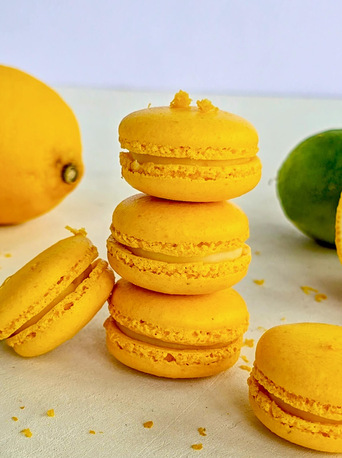 Une pile verticale de trois macarons jaunes au citron avec une collerette parfaite, entourée de citrons frais coupés, de zestes et de feuilles de basilic sur un fond blanc lumineux.