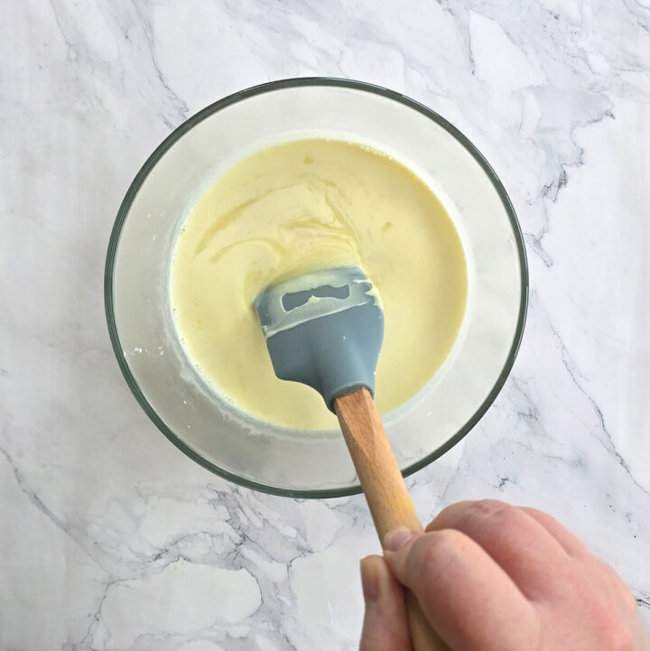 Une main mélangeant une préparation liquide d'un jaune crémeux et onctueux dans un bol en verre transparent, à l'aide d'une spatule en silicone grise à manche en bois. Le bol repose sur une surface en marbre blanc.