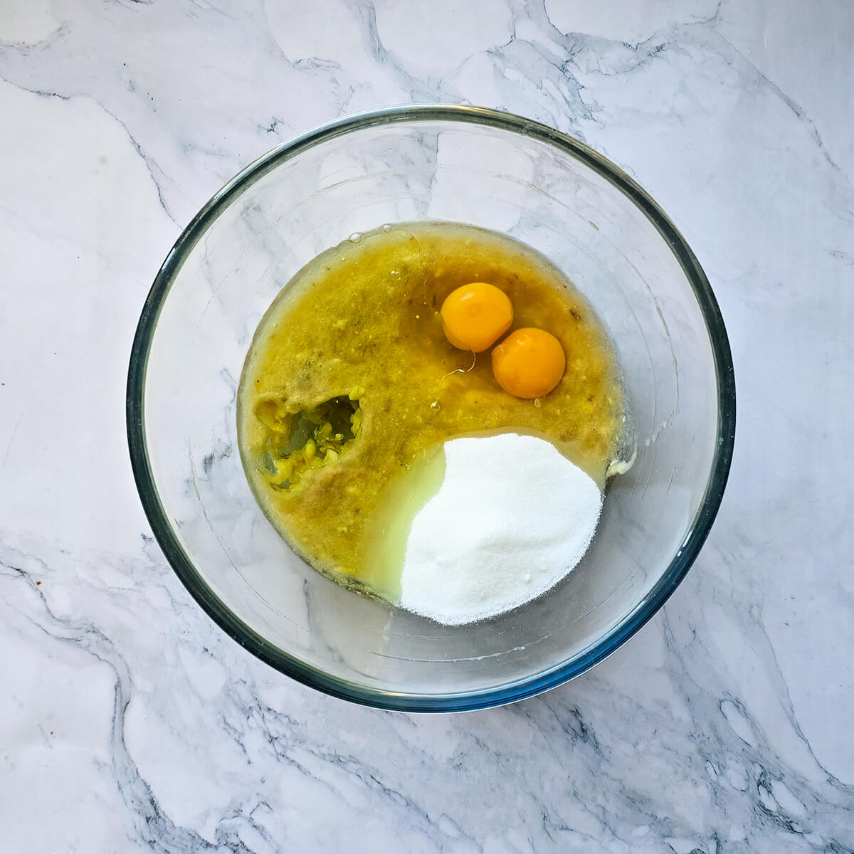 Vue de dessus d'un bol en verre contenant de la purée de banane, deux jaunes d'œufs entiers et de sucre blanc, prêts à être mélangés sur un plan de travail en marbre.