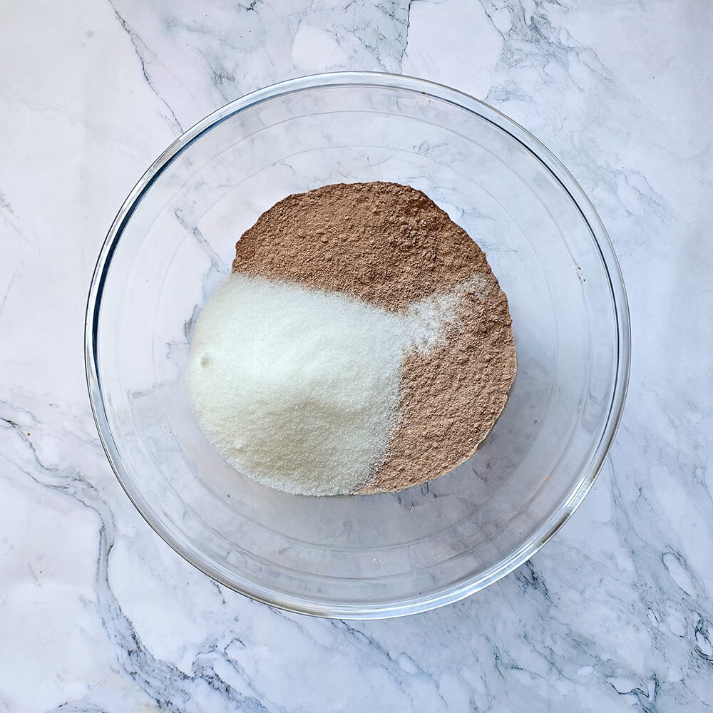 Gros plan des ingrédients secs (farine, sucre blanc et cacao en poudre) mesurés et disposés dans un saladier en verre, prêts à être mélangés pour la base du chiffon cake.