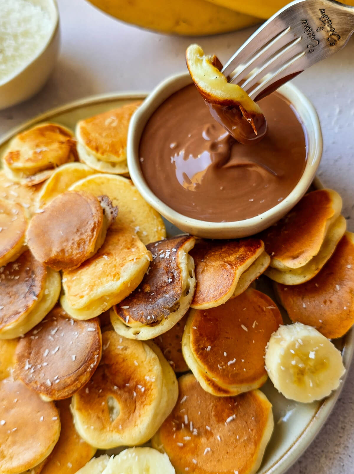 Une fourchette avec la marque "Cuisine & Création" gravée sur le manche prélève un mini-pancake doré à la banane et le trempe généreusement dans un ramequin rempli de pâte à tartiner au chocolat onctueuse, au-dessus de l'assiette de "pancake cereal".