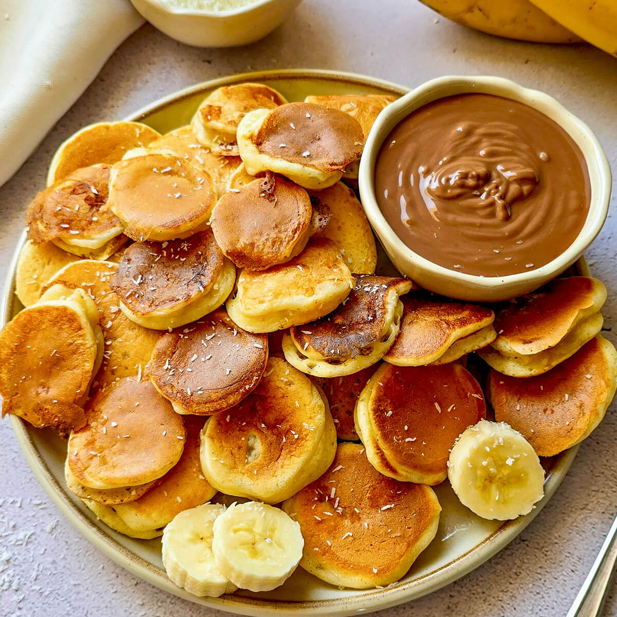 Une assiette remplie de "pancake cereal" (mini-billes de pancakes healthy à la banane) dorés et moelleux, parsemés de noix de coco râpée. Au centre, un bol de pâte à tartiner au chocolat pour le dip, avec des rondelles de banane fraîche et des pépites de chocolat en garniture.
