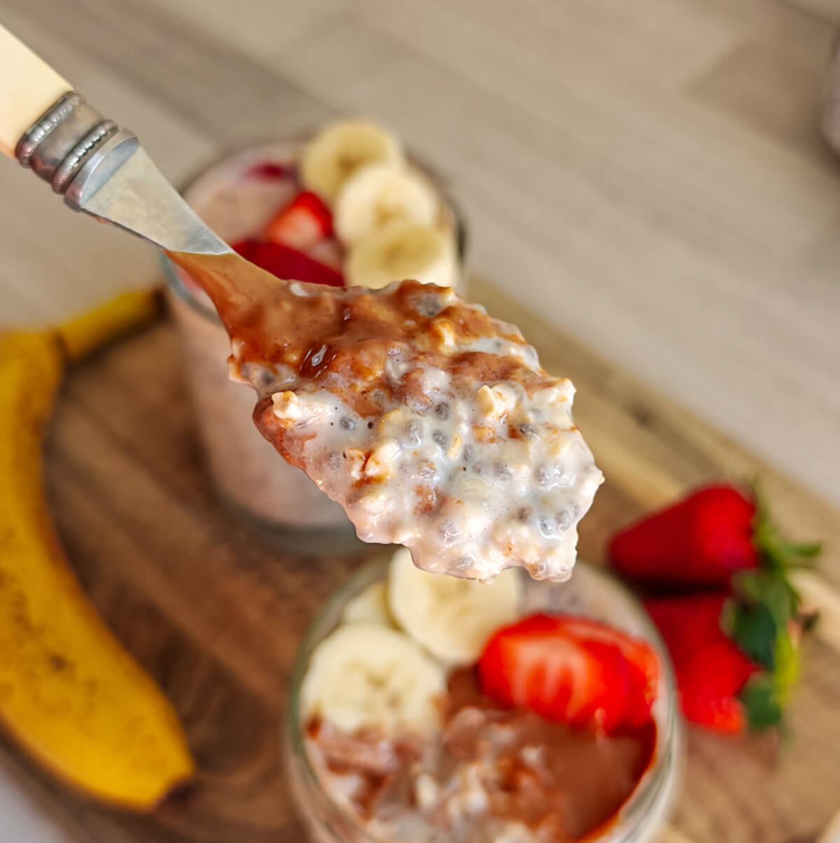 Gros plan sur une cuillère soulevant une bouchée d'overnight oats crémeux aux graines de chia et beurre d'amande, avec des bocaux garnis de bananes et de fraises en arrière-plan flou.