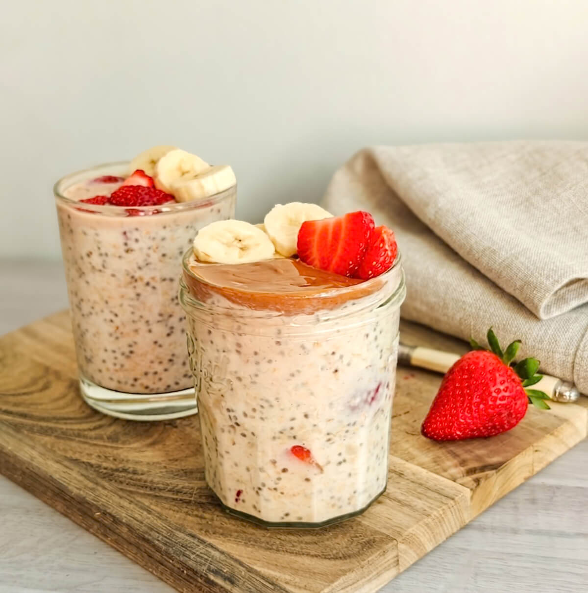 Deux bocaux d'overnight oats fraise banane et graines de chia, garnis de rondelles de banane, de fraises fraîches et d'une couche onctueuse de beurre d'amande, posés sur une planche en bois avec un torchon en lin en arrière-plan.