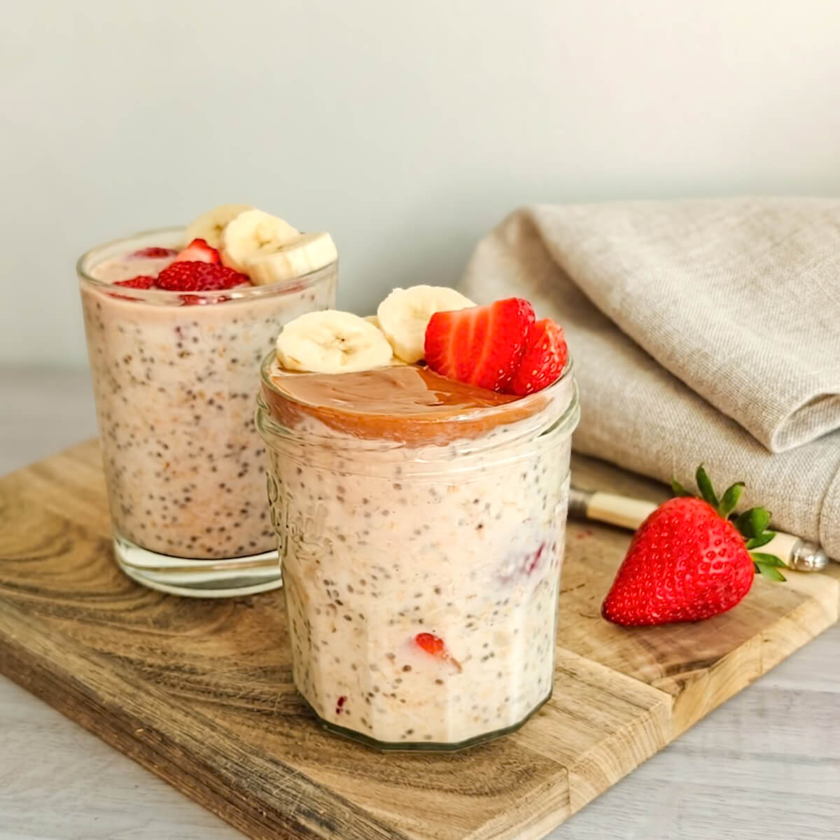 Deux bocaux d'overnight oats fraise banane et graines de chia, garnis de rondelles de banane, de fraises fraîches et d'une couche onctueuse de beurre d'amande, posés sur une planche en bois avec un torchon en lin en arrière-plan.