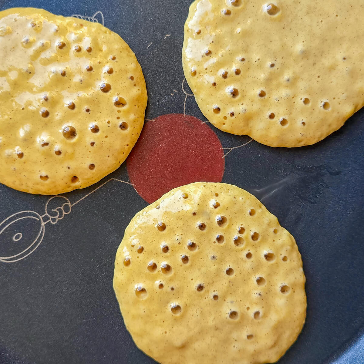 Trois pancakes ronds et moelleux, de couleur brun doré, cuisent sur une poêle antiadhésive T-fal noire. La pâte commence à faire des bulles à la surface.